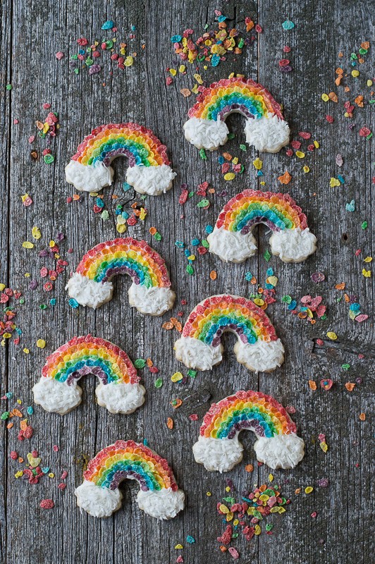 Eight Rainbow Sugar Cookies covered in fruity pebbles and shredded coconut on a wooden table.