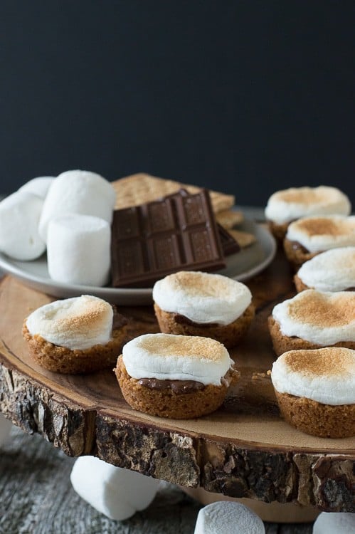 smores bites on wood cake stand with marshmallows, chocolate and graham crackers in background