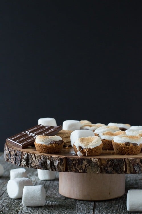 s'mores bites on round wood cake stand with marshmallows in foreground on dark background