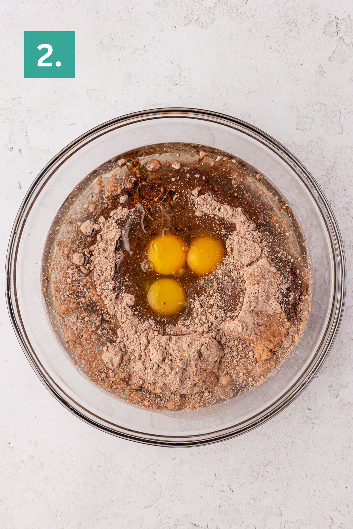A glass mixing bowl with chocolate cake mix, three raw eggs, oil, and water on a light surface. A green square labeled 2. is in the top left corner, showing step two in this delicious earthquake cake recipe.
