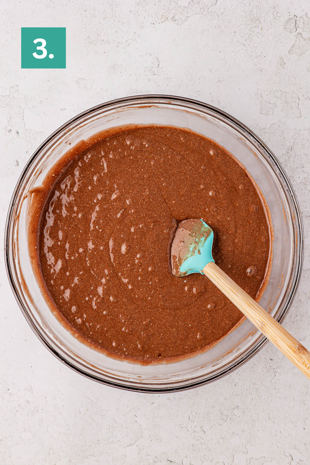 A glass bowl filled with rich earthquake cake batter is shown with a blue spatula inside. The bowl sits on a light countertop, while a green box with the number 3 appears in the upper left corner.