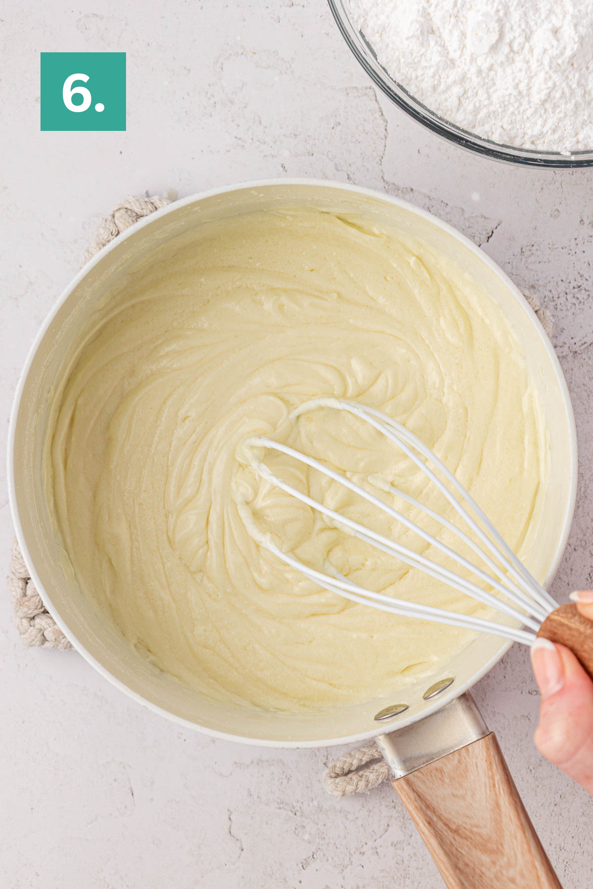 A hand whisks a creamy, pale earthquake cake batter in a light saucepan, with a bowl of flour in the background. A green box labeled 6 appears in the top left corner, marking this recipe step.