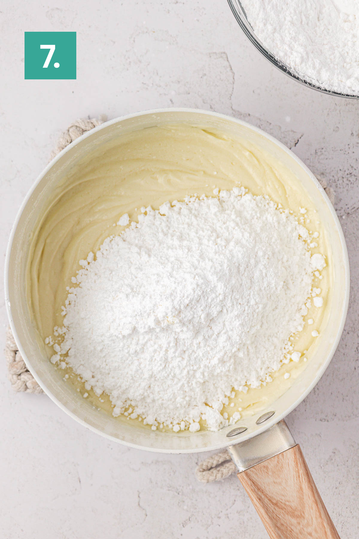 A saucepan filled with creamy earthquake cake batter topped with a mound of powdered sugar sits on a white surface. A 7. label is in the top left corner, and a bowl with more powdered sugar is nearby.