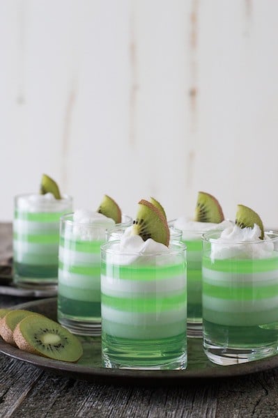 Four Layered Melon Kiwi Jello Cups garnished with fresh kiwis on a silver platter.