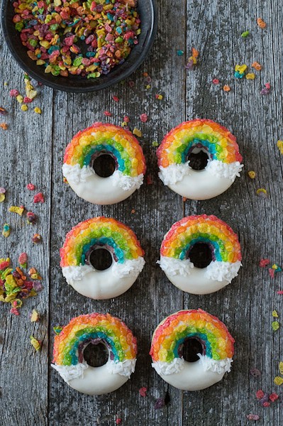 Six Rainbow Donuts surrounded by fruity pebbles on a wooden table.