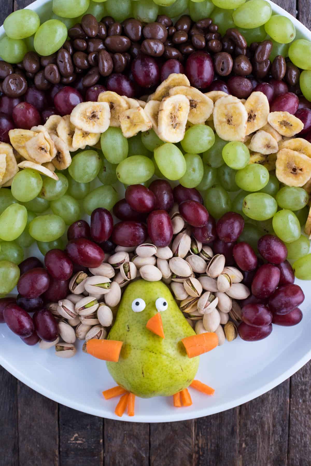 turkey shaped fruit tray with pistachios, grapes, banana chips, and pear and carrots on round white platter on wood background
