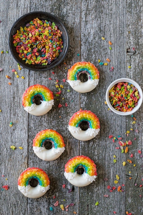 Six Rainbow Donuts next to two bowls of fruity pebbles on a wooden table.