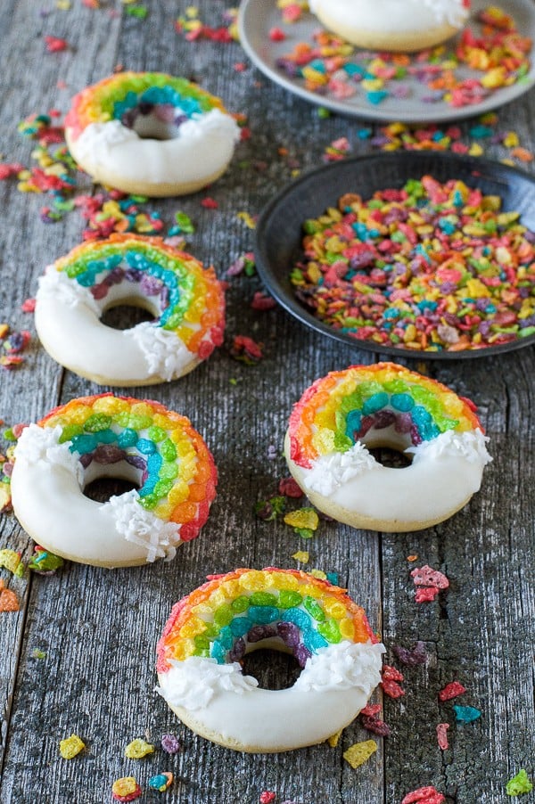 Five homemade Rainbow Donuts covered in white chocolate, coconut shavings and fruity pebbles on a wooden table.