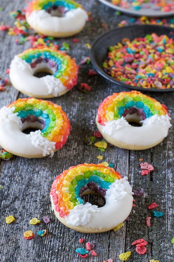 Four easy Rainbow Donuts surrounded by colorful fruity pebbles on a wooden table.