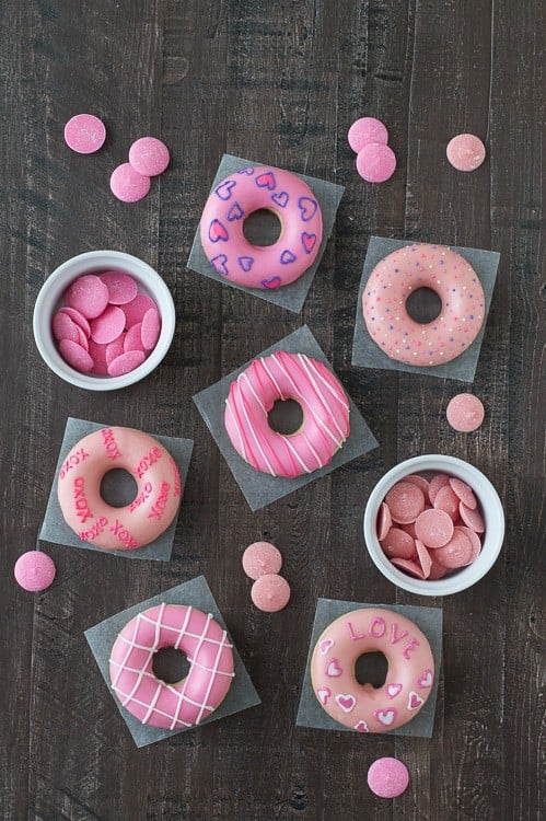 Six delicious Valentine’s Day Donuts and two white bowls with two different shades of pink candy melts on a wooden table. 