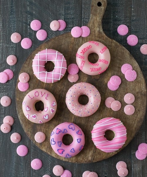Six Valentine’s Day Donuts on a wooden board surrounded by pink candy melts.