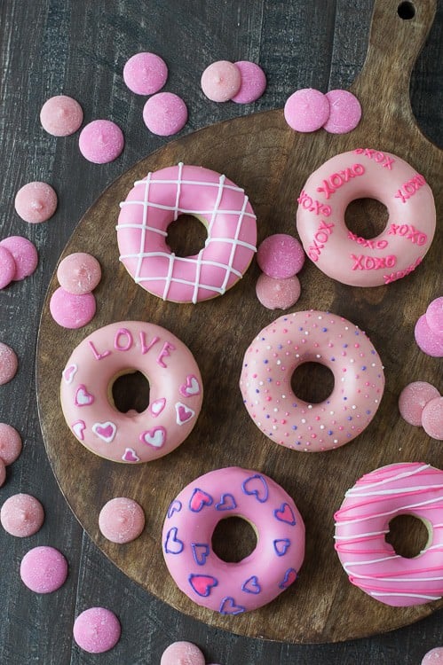Six pink Valentine’s Day Donuts surrounded by pink candy melts on a wooden board on a wooden table. 