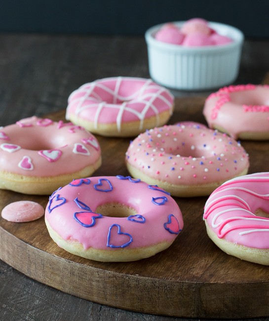 Six easy and delicious Valentine’s Day Donuts on a wooden board with a small white bowl of pink candle melts next to it. 