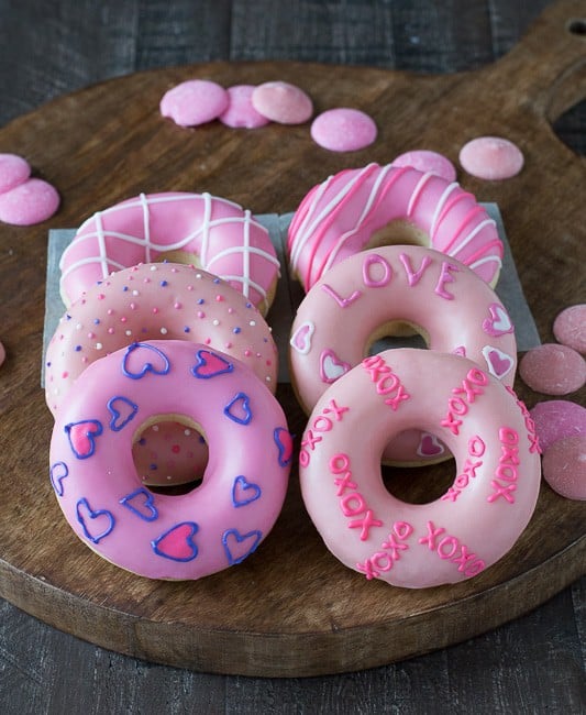 Six Valentine’s Day Donuts covered in pink frosting on a wooden board. 