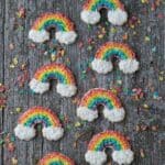 Eight Rainbow Sugar Cookies decorated with Fruity Pebbles and coconut shavings on a wooden table.