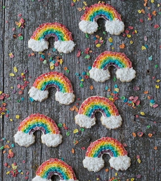 Eight Rainbow Sugar Cookies decorated with Fruity Pebbles and coconut shavings on a wooden table.