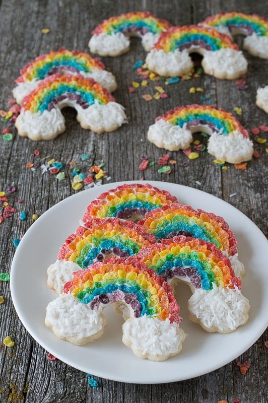 Five Rainbow Sugar Cookies on a white serving plate with six more rainbow sugar cookie treats in the background on a wooden table.