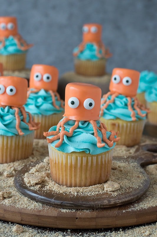 Seven easy vanilla Octopus Cupcakes covered in blue buttercream frosting on a round wooden board.