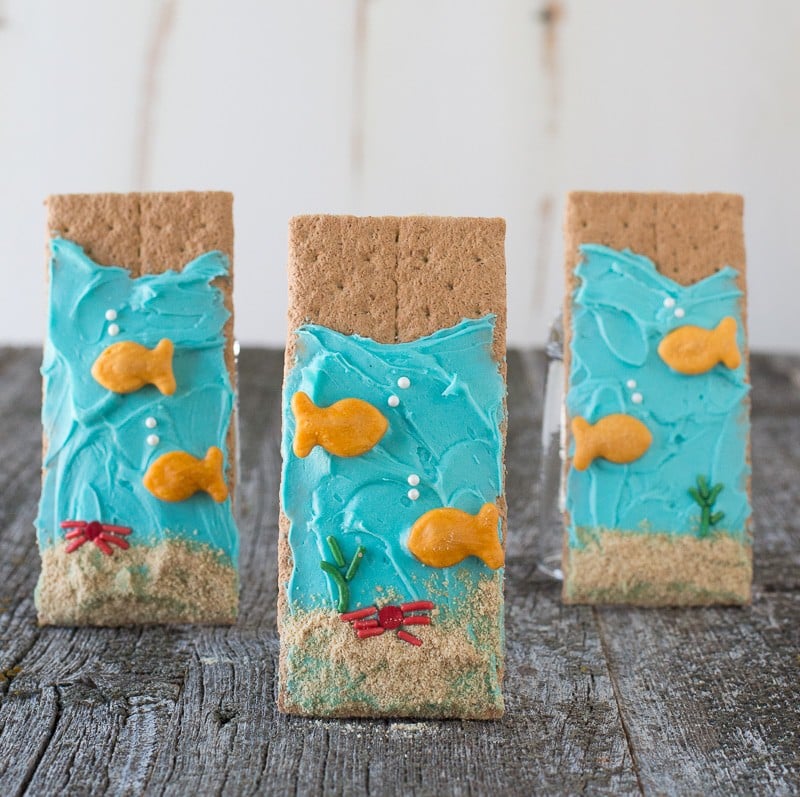 Three delicious Under the Sea Graham Crackers decorated with blue buttercream frosting and goldfish crackers standing on a wooden table.