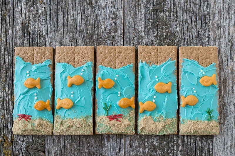 Five Under the Sea Graham Crackers with blue buttercream and goldfish crackers on a wooden table. 