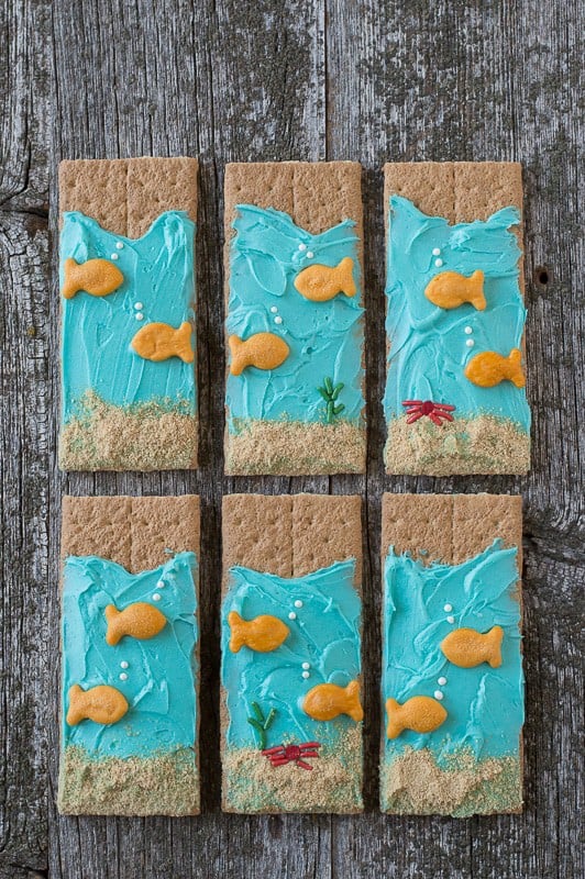 Six Under the Sea Graham Crackers with blue buttercream and goldfish crackers on a wooden table. 