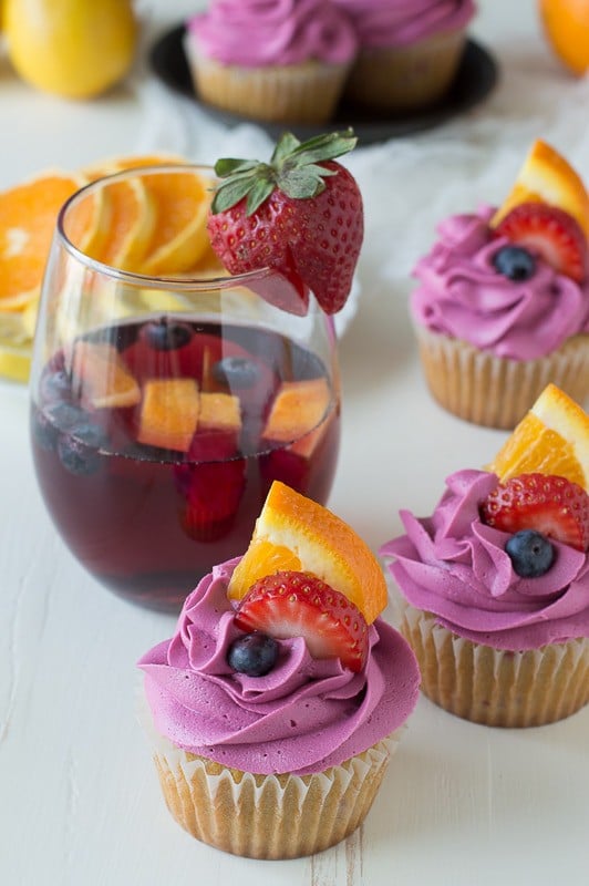 Two homemade Sangria Cupcakes with red wine buttercream garnished with blueberries, slice orange and strawberries next to a glass of sangria.