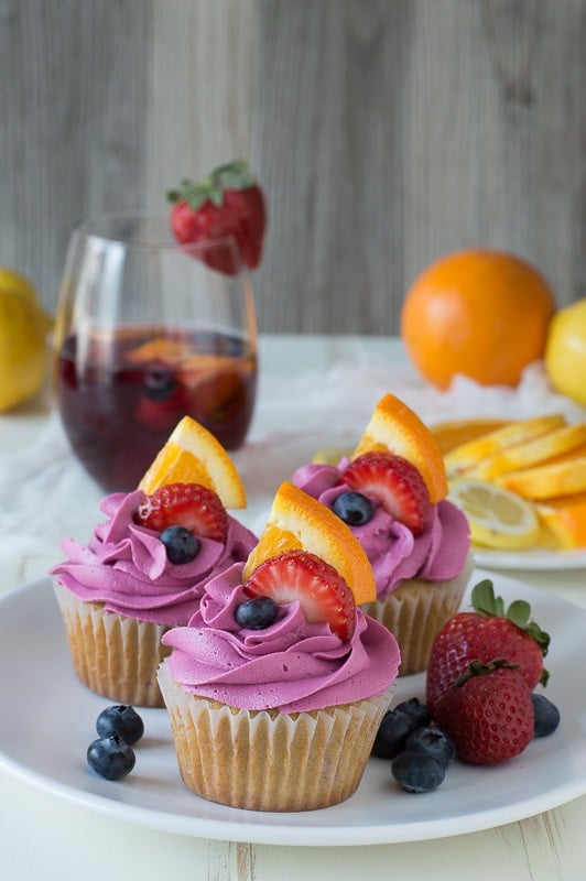 Three Sangria Cupcakes with pink frosting surrounded by fresh strawberries and blueberries on a white serving plate.