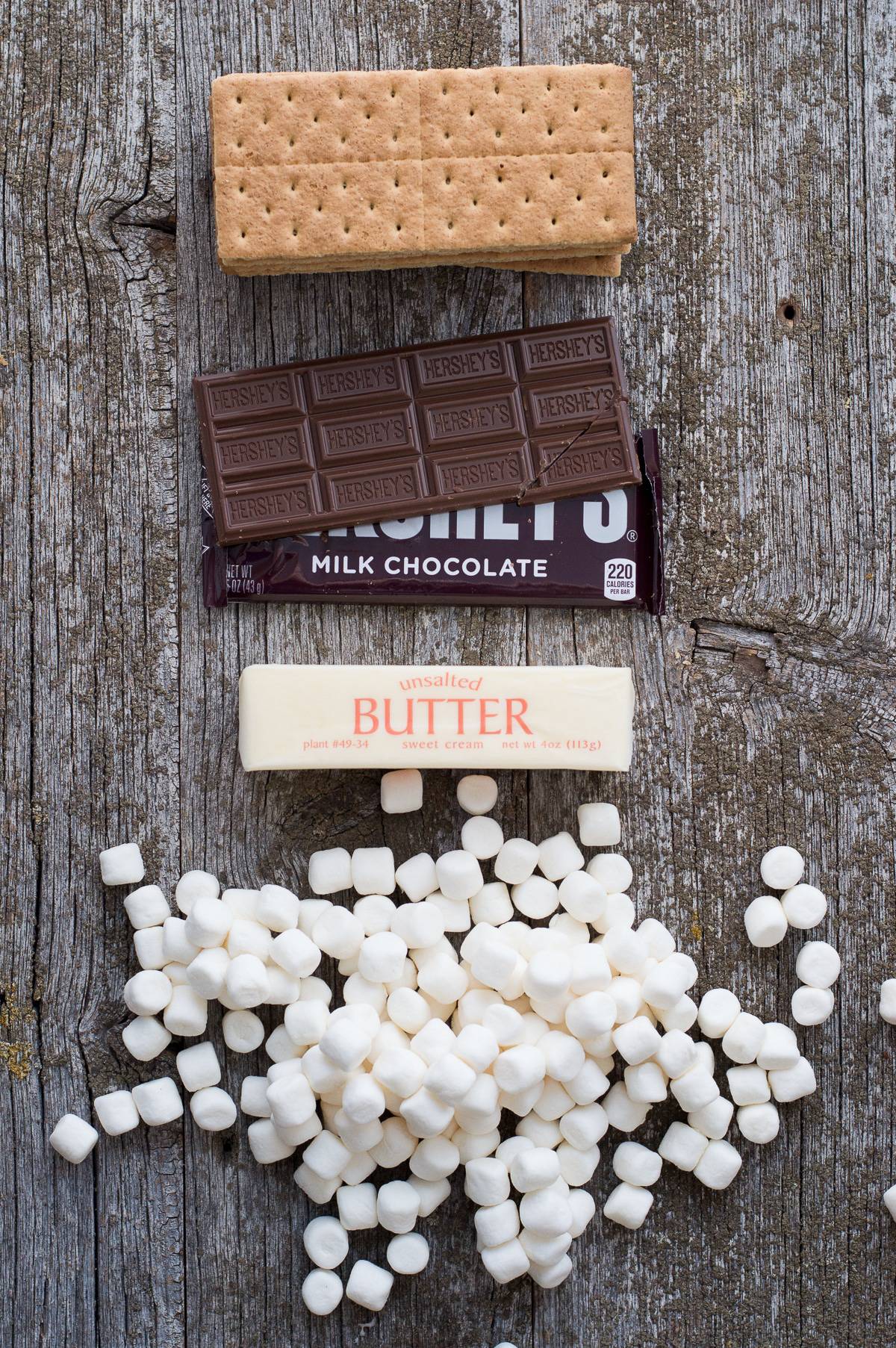 graham crackers, hershey's chocolate bars, stick of butter and mini marshmallows on gray wood background