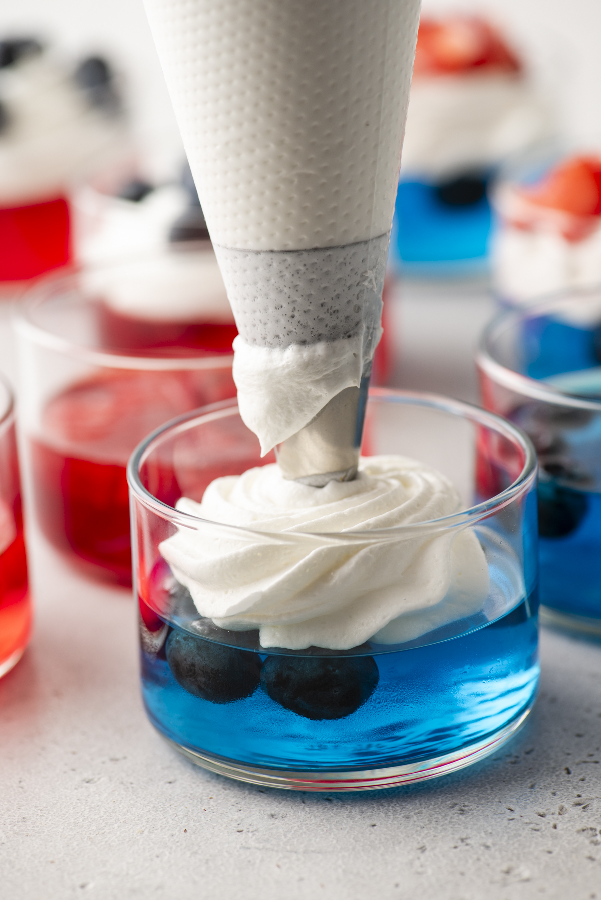 A close-up of a piping bag adding whipped cream atop blue gelatin dessert with blueberries in a glass cup&mdash;perfect 4th of July jello cups. Red gelatin desserts with whipped cream are visible in the background.