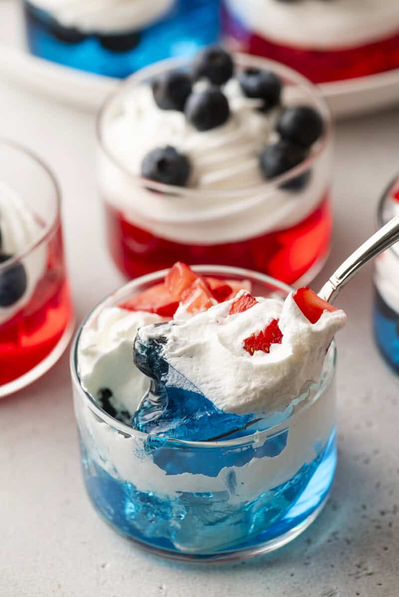 A glass cup filled with blue gelatin, whipped cream, and chopped strawberries&mdash;perfect 4th of July Jello cups&mdash;sits in front of similar desserts with whipped cream and blueberries. A spoon scoops up a festive bite.