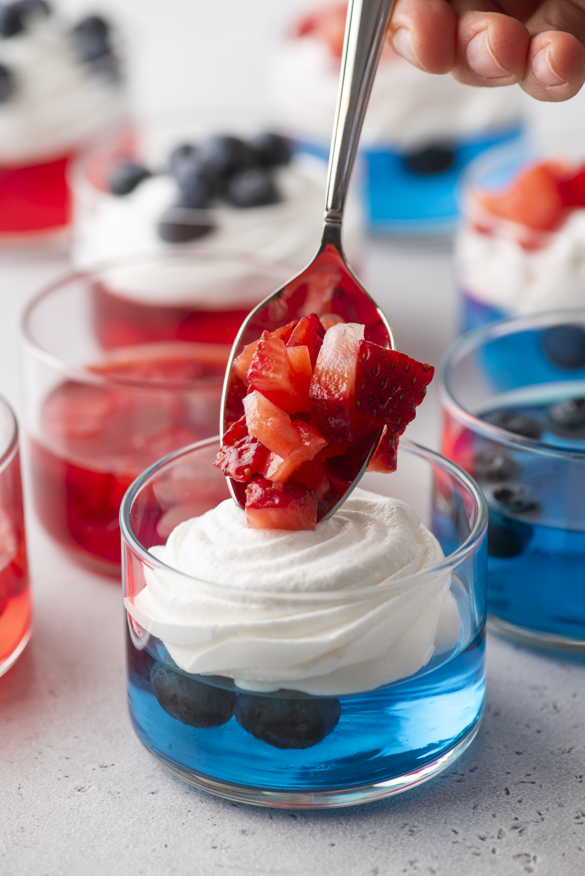 A spoonful of chopped strawberries is being placed onto a 4th of July jello cup with blue jelly, blueberries, and whipped cream in a glass. More colorful jelly desserts are visible in the background.
