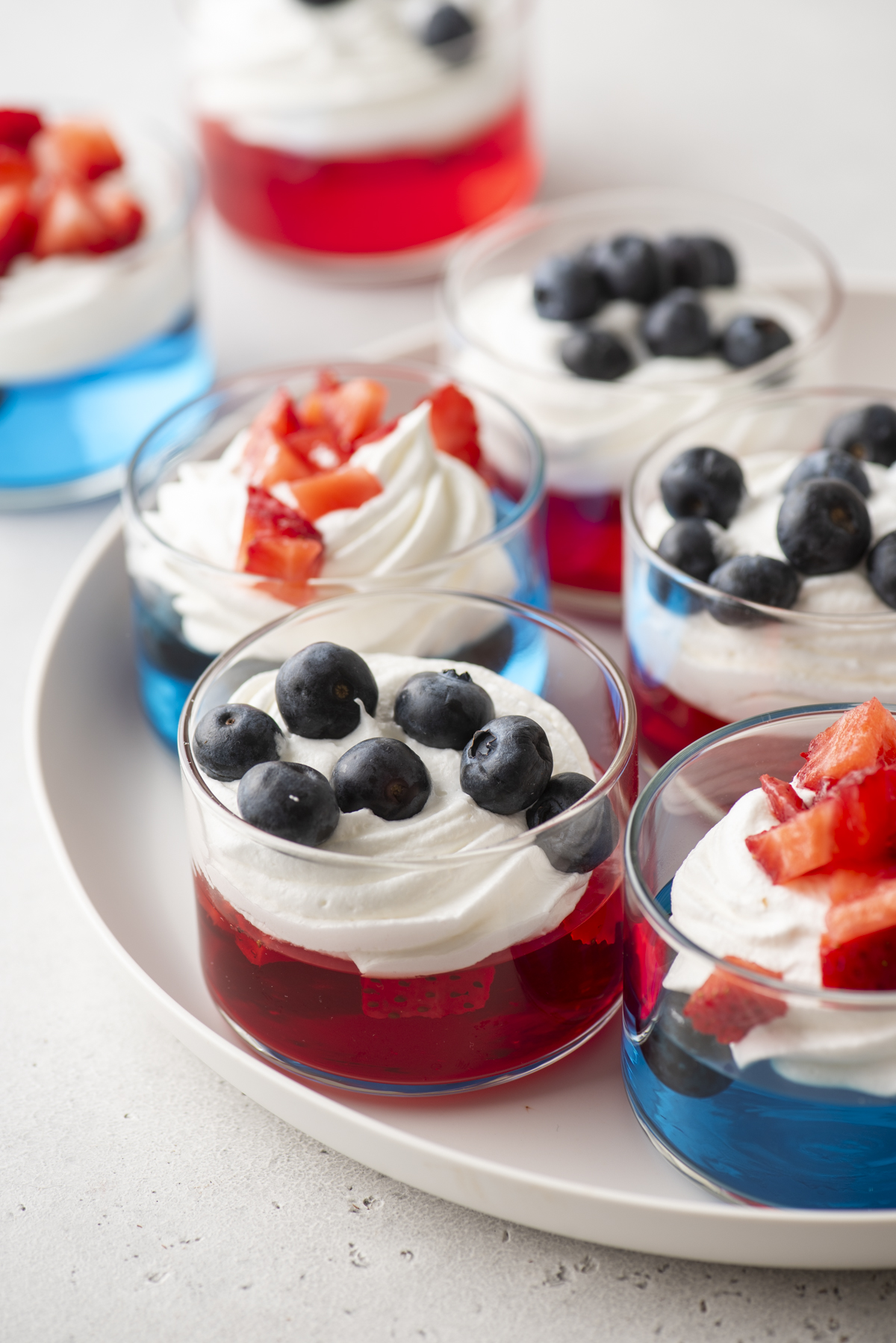 Small glass cups filled with red and blue gelatin, topped with whipped cream and fresh blueberries or chopped strawberries, make festive 4th of July jello cups arranged beautifully on a white plate.