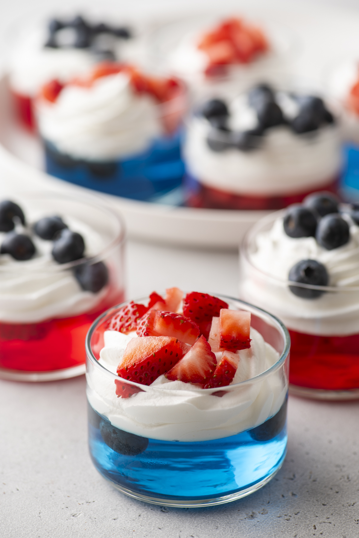 Several 4th of July jello cups filled with red and blue gelatin, topped with whipped cream and fresh strawberries or blueberries, are arranged on a light surface. The desserts look festive and colorful.