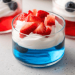 A glass cup filled with blue gelatin, topped with whipped cream, fresh strawberry pieces, and blueberries—perfect for festive 4th of July jello cups. Another similar dessert is blurred in the background.