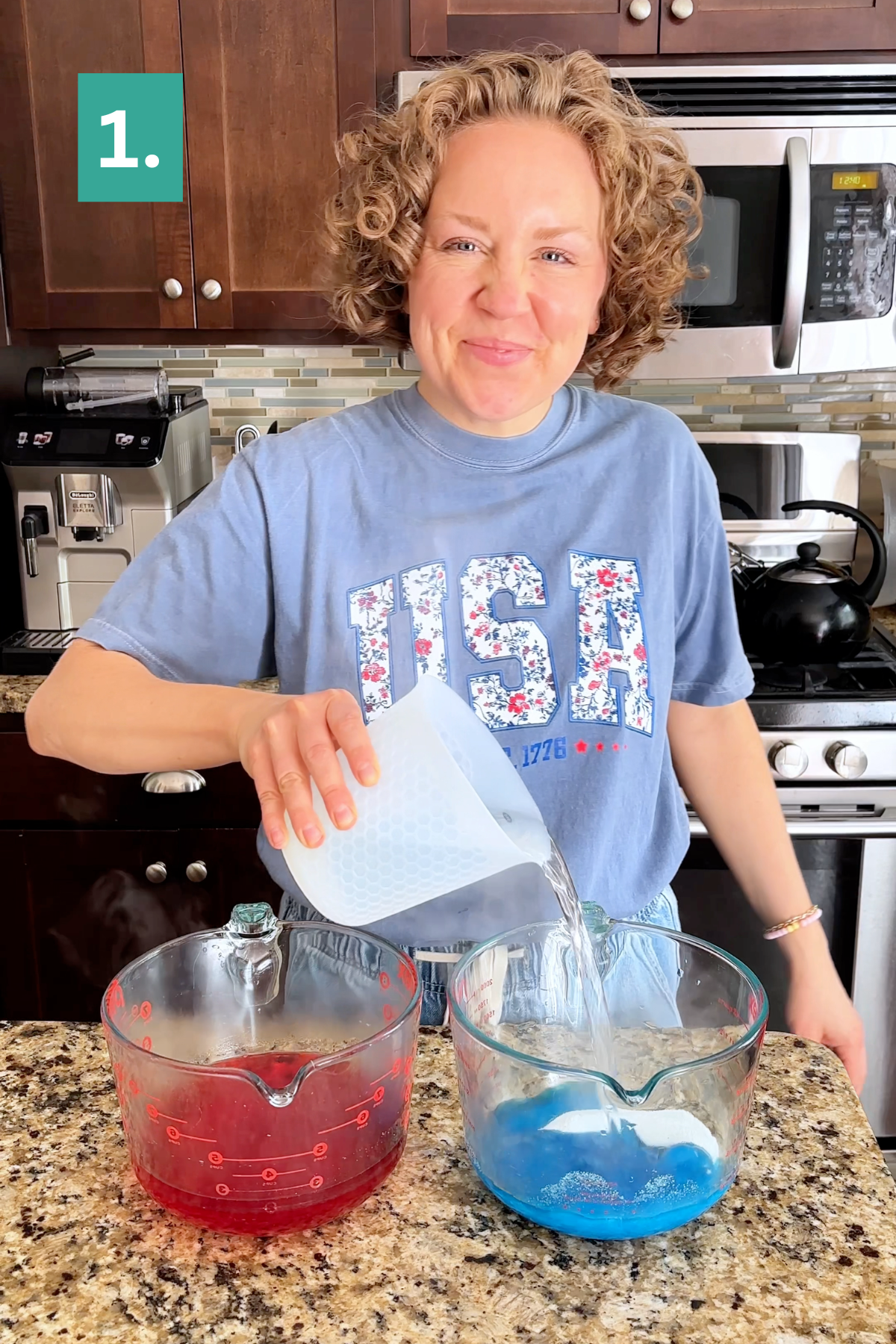 A smiling woman in a blue USA shirt pours liquid from a white measuring cup into a glass bowl with blue liquid, next to a bowl with red liquid, as she prepares 4th of July jello cups in her kitchen. A green box with the number 1 is in the top left.