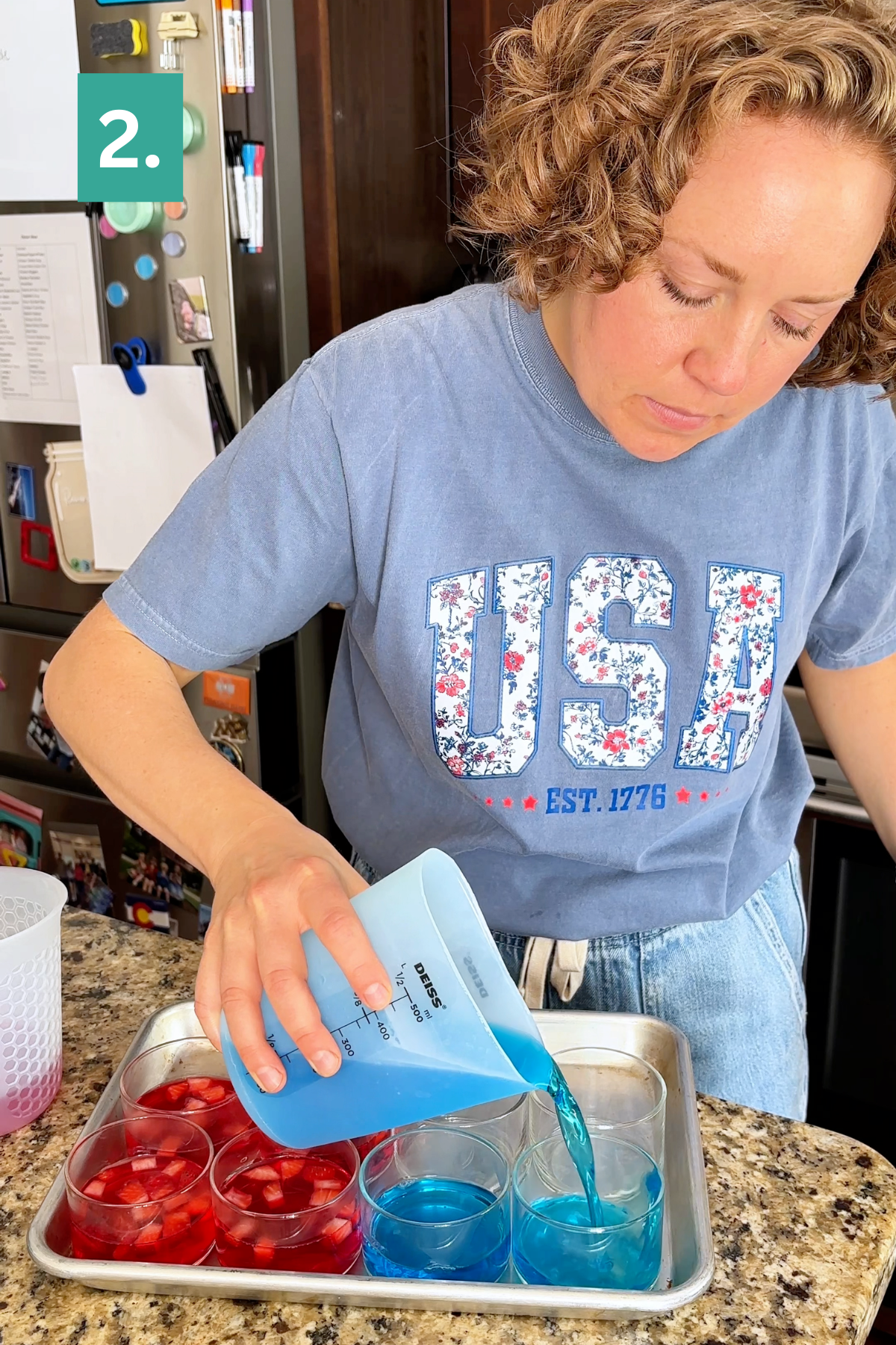 A woman wearing a blue USA t-shirt pours blue liquid into clear cups filled with red fruit, creating festive 4th of July jello cups on a metal tray on the kitchen counter. A 2. label appears in the corner.