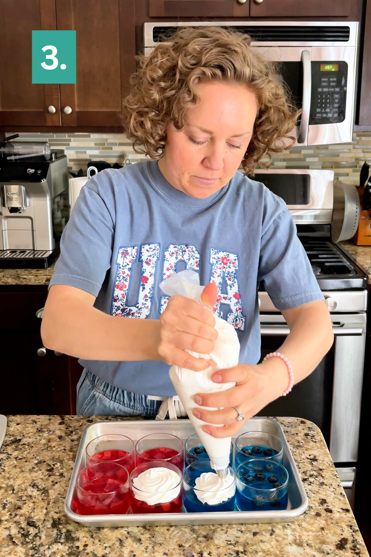 A woman in a blue &ldquo;USA&rdquo; shirt pipes white whipped topping onto 4th of July Jello cups layered with red and blue desserts in clear cups on a kitchen counter. A &ldquo;3.&rdquo; label appears in the top left corner, marking this step in the process.
