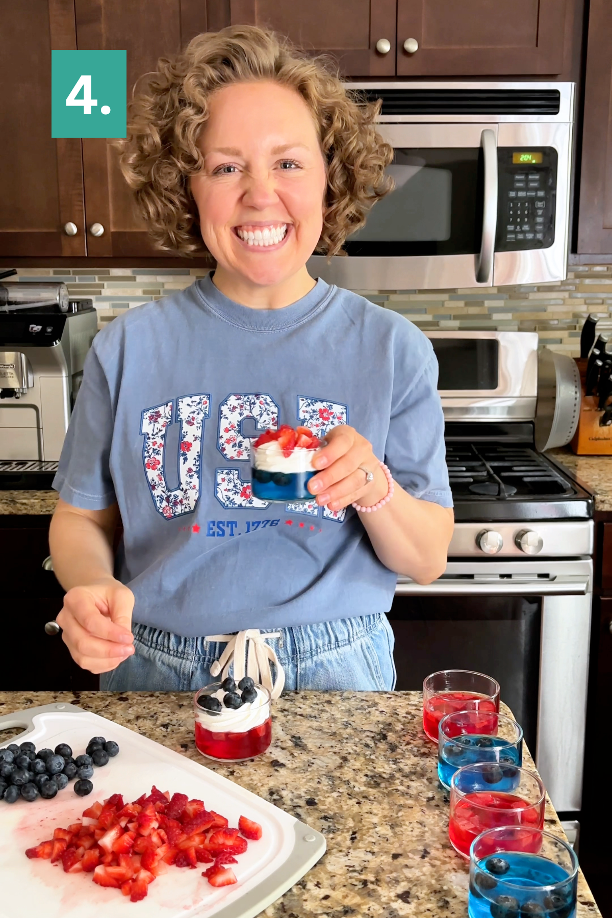 A smiling woman in a USA shirt stands in a kitchen, holding layered 4th of July jello cups with strawberries and blueberries. On the counter are chopped strawberries, whole blueberries, and other patriotic desserts. A green box with 4. is in the corner.