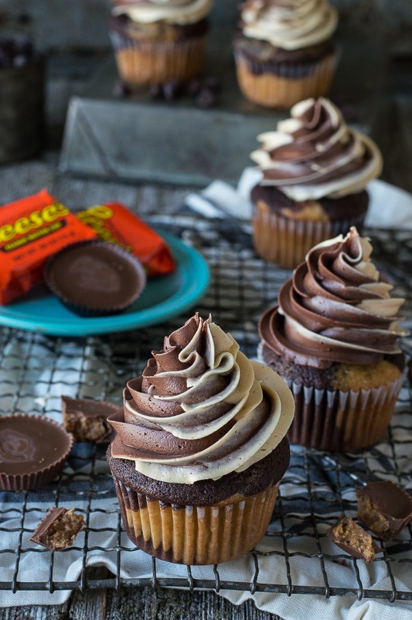Reese's Cupcakes - Tis the season to use up that halloween candy. And while eating it straight out of the wrapper is probably my favorite way.. incorporating that candy into recipes is a good way to impress your friends and coworkers. Three of the ultimate Reese’s Cupcakes with peanut butter buttercream frosting with peanut butter cups on a blue plate on a cooling rack.