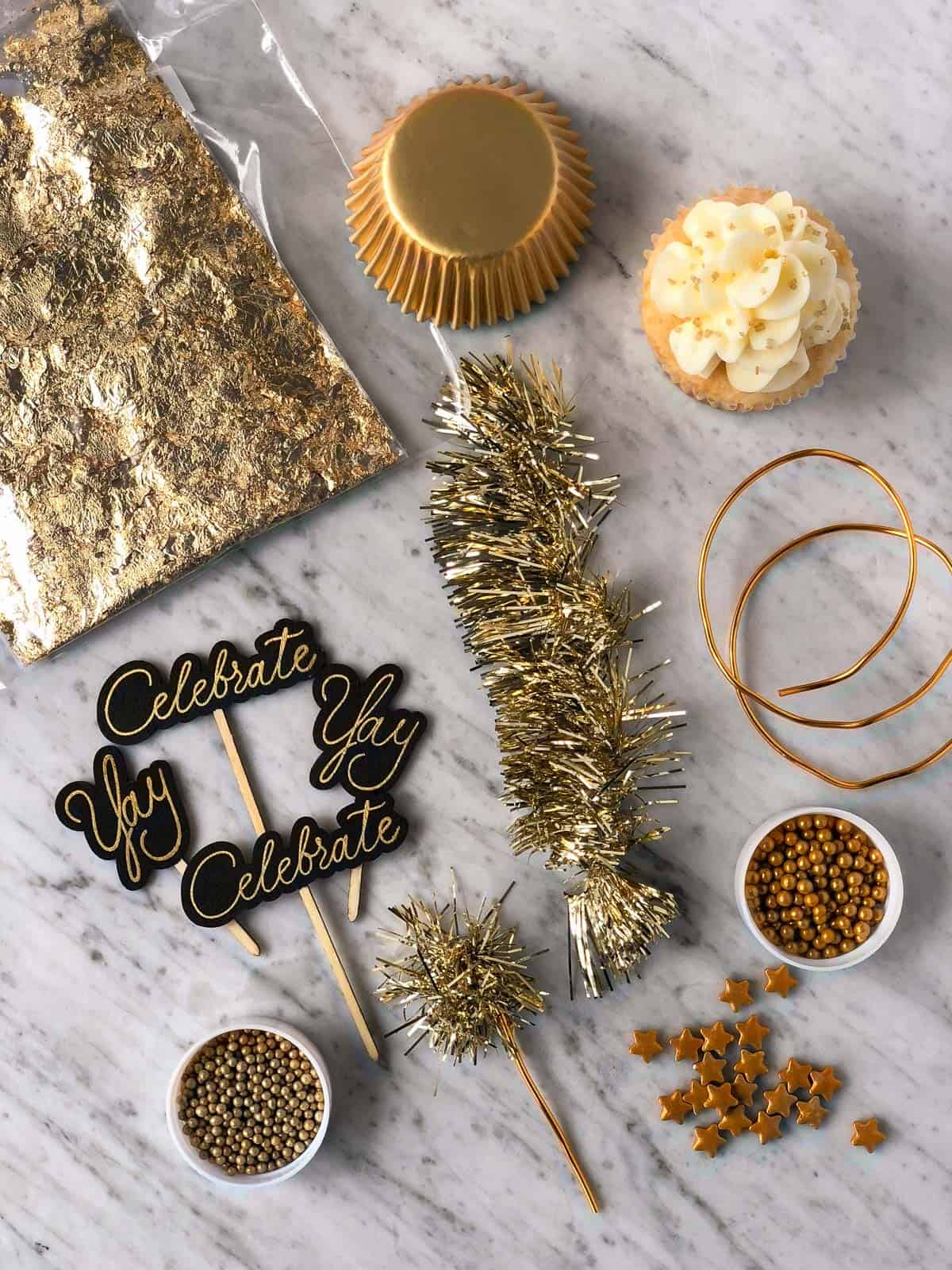 supplies used to decorate new year's eve cupcakes including gold sprinkles, gold stars, gold muffin liners, edible gold leaf, celebrate cupcake toppers, and gold tinsel