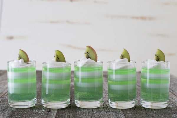 Five Layered Melon Kiwi Jello Cups garnished with kiwis on a wooden table.