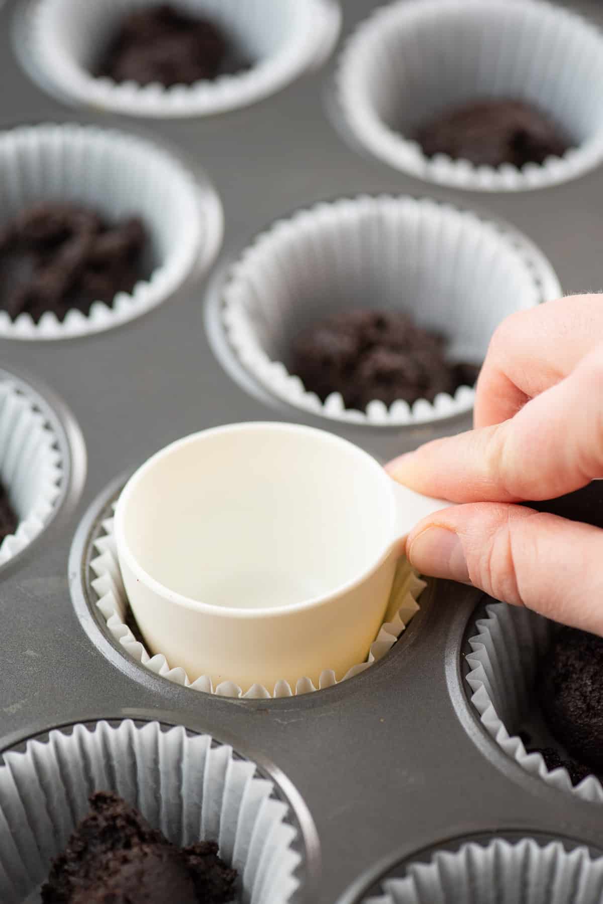 white measuring cup packing down oreo crust in metal muffin pan