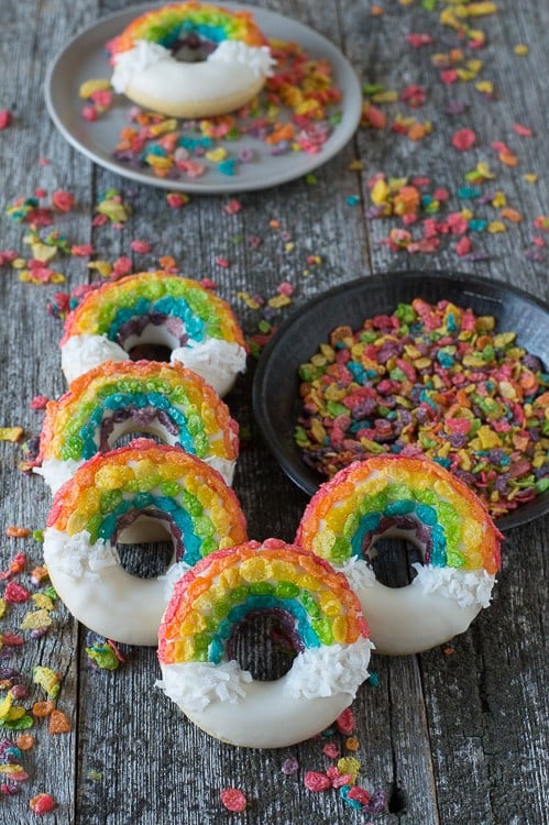 Five Rainbow Donuts next to a bowl of fruity pebbles on a wooden table.