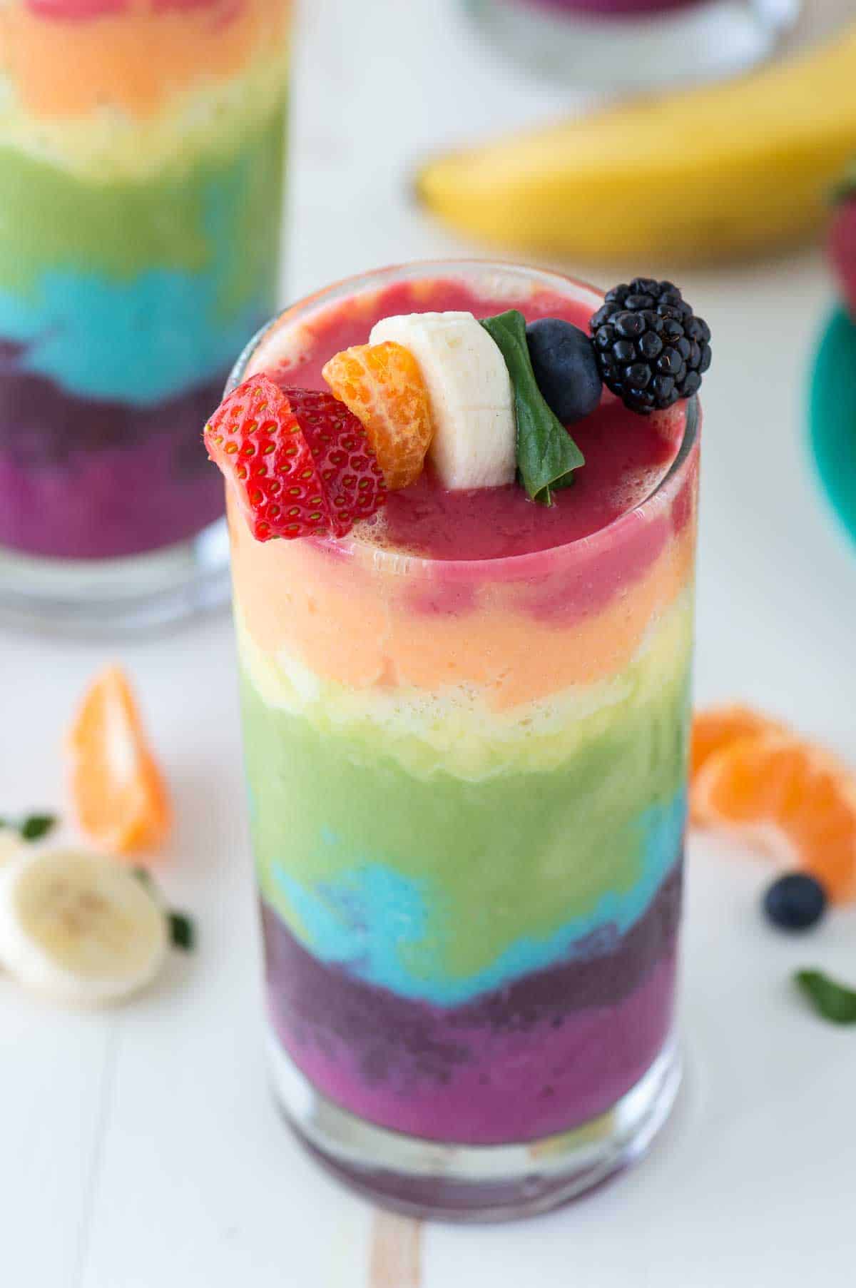 rainbow smoothie layered in glass cup with strawberry, orange, banana, spinach, blueberry and blackberry on top