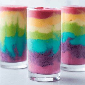 Three glasses, filled with vibrant, layered rainbow smoothies, display a stunning gradient—from pink at the bottom to purple, blue, green, yellow, and red atop. This colorful array forms an exquisite visual symphony on the white surface.