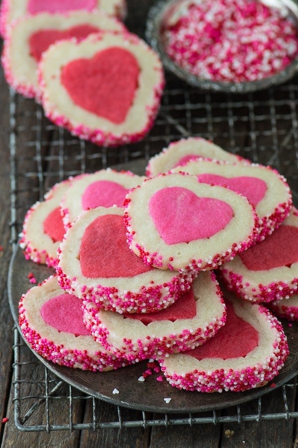 Stack of delicious Homemade Valentine’s Day cookies on a cooling rack next to a bowl of pink and white sprinkles. 