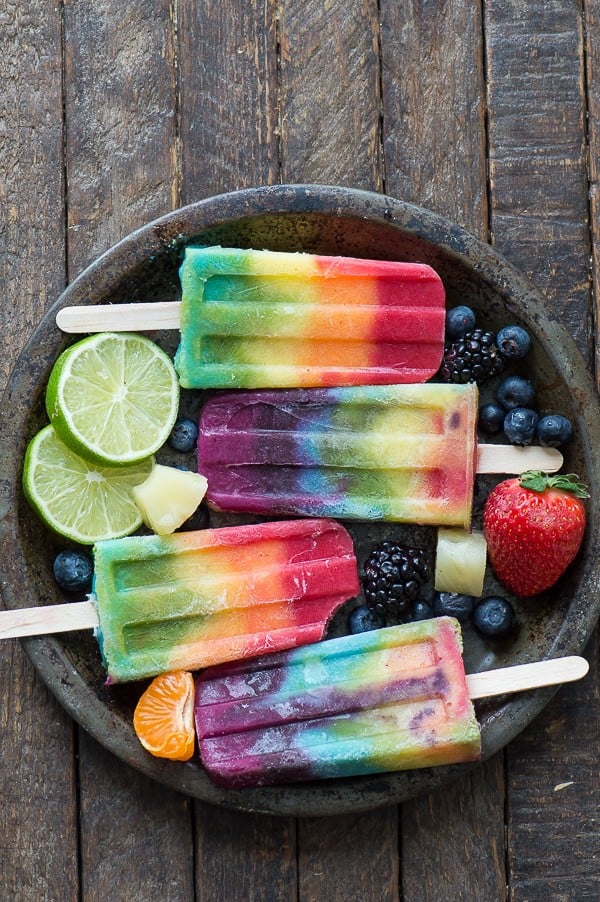 rainbow popsicles on metal plate with blueberries, limes, strawberry, orange surrounding the popsicles