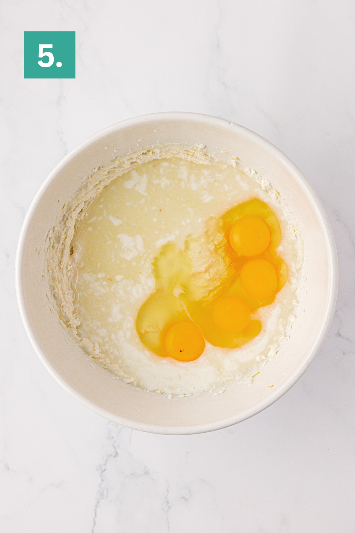 A white mixing bowl containing three cracked eggs, runny egg whites, and creamy 9x13 Lemon Cake batter sits on a white marble surface. A green square with the number 5 is in the top left corner.