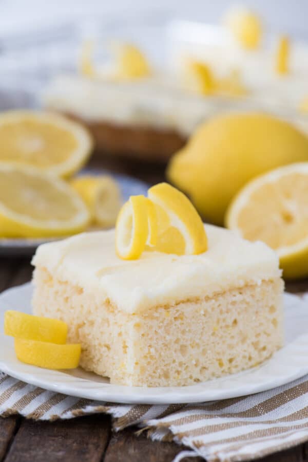 piece of lemon cake with lemon frosting on white plate with lemons in the background