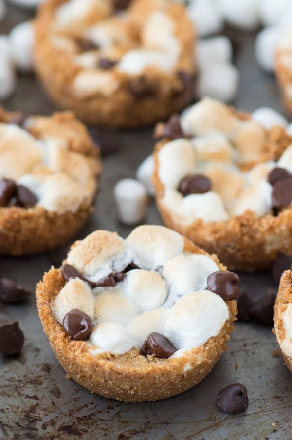 5 ingredient indoor S’mores Cups! Make these in a regular muffin pan. Like a cross between s'mores cookies and s'mores cupcakes! Arrangement of four 5 ingredient indoor S’mores Cups on a baking sheet.